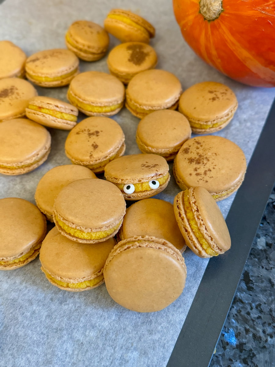 a pile of pumpkin macarons with spices on a baking tray, one with eyes stuck to the ganache filling