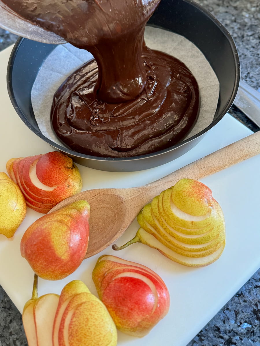 pouring chocolate cake batter into a lined cake tin next to sliced pears