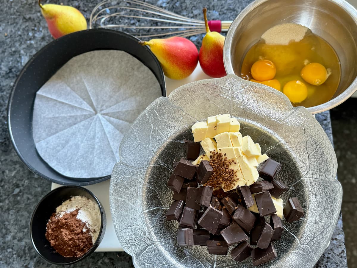 a bowl of chocolate pieces, butter and coffee and another with eggs and sugar next to a lined cake tin and 3 fresh pears