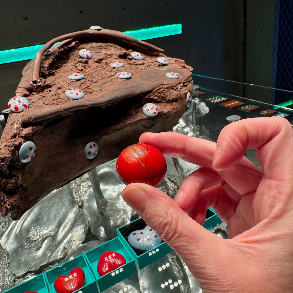 holding a red demi sphere red shiny chocolate in front of chocolate hearts and a large chocolate sculpture in a Parisian shop