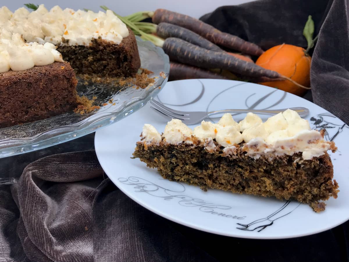 slice of dark cake topped with cream cheese frosting on a plate with purple carrots and orange in background