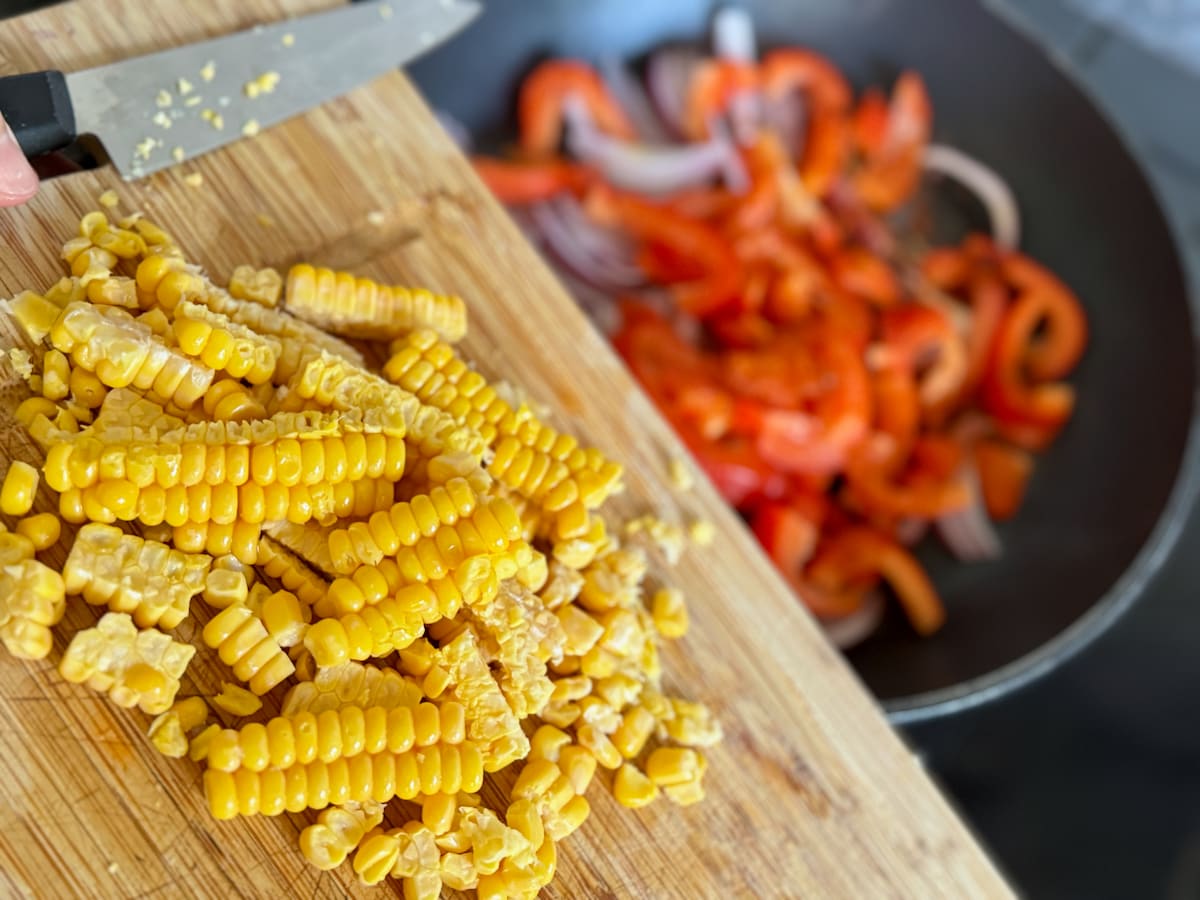 adding fresh corn to a frying pan of chopped red bell pepper and onion