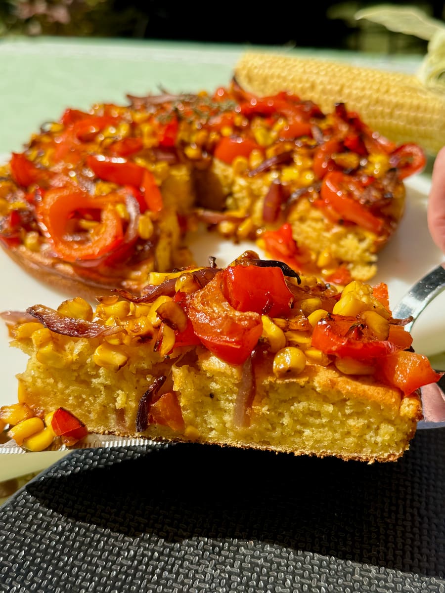 cutting a slice of cheesy cornbread, light and moist topped with corn and other roasted vegetables