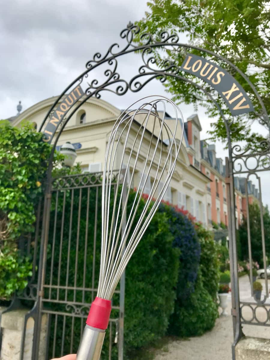 holding a whisk in front of a large hotel gate inscribed with the words birthplace of Louis XIV in French