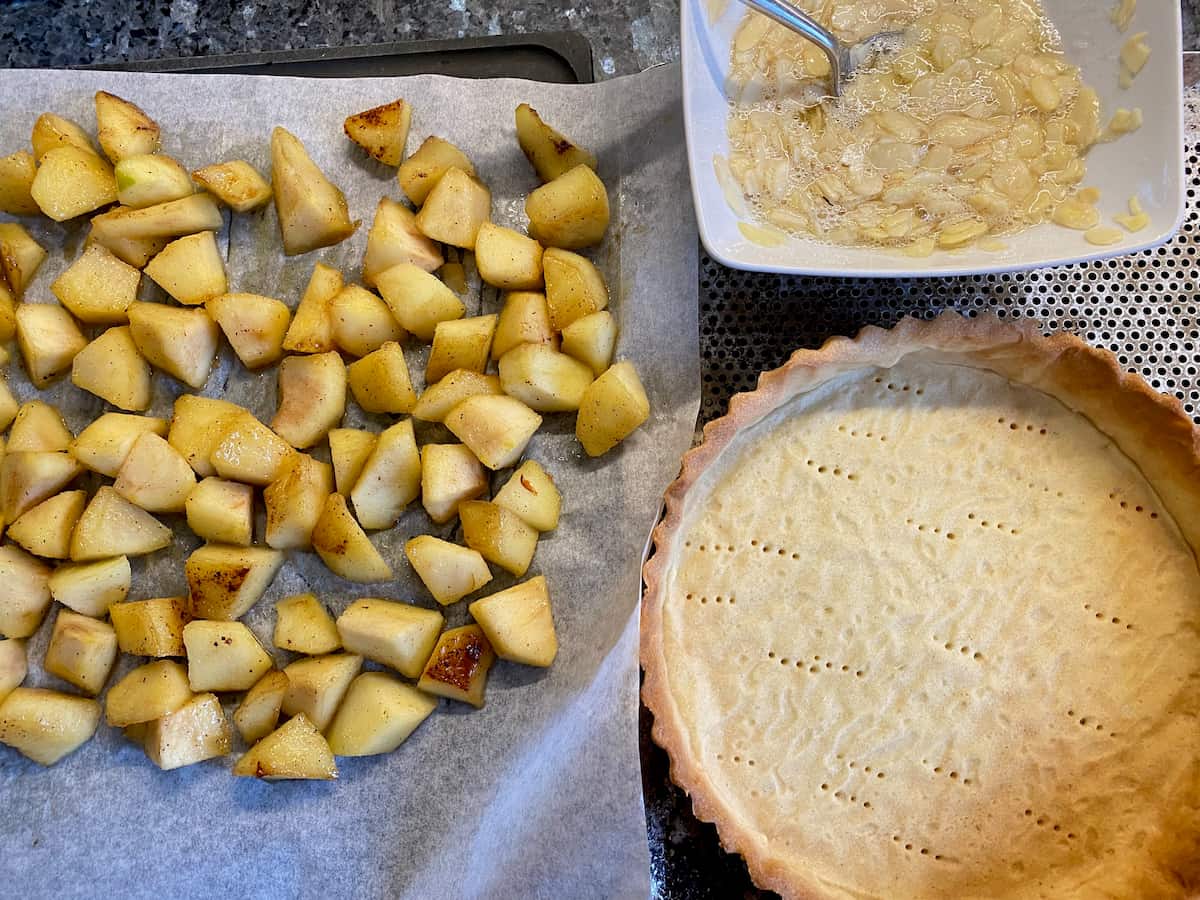 empty baked tart shell with cooked apple chunks on parchment and a bowl of egg whites with almonds and sugar