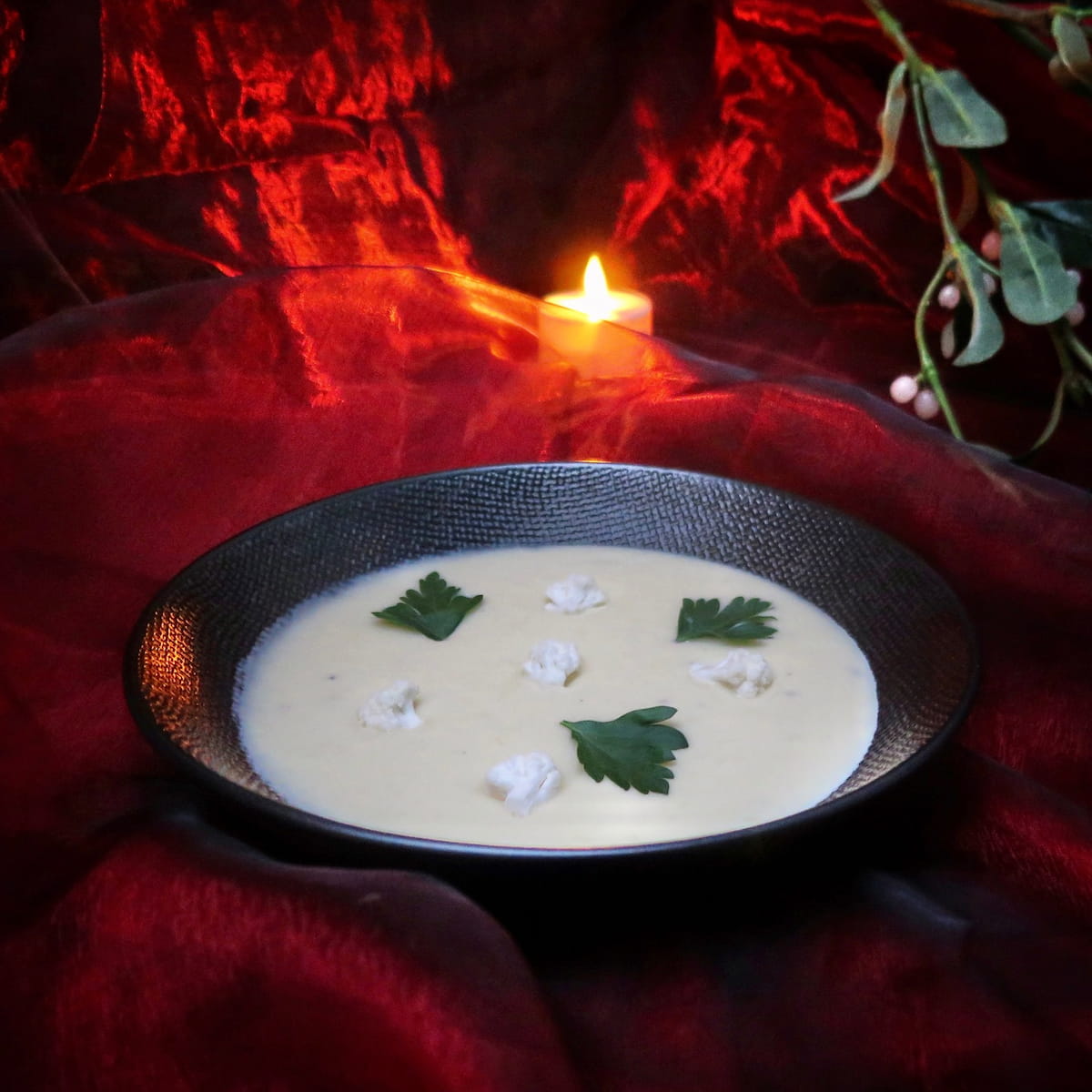 black bowl of creamy soup with cauliflower florets on silk red, mistletoe and candle lit
