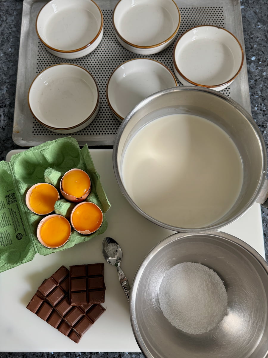 4 egg yolks, milk chocolate, sugar and a pan of half and half milk and cream next to a baking tray of 5 shallow ramekins