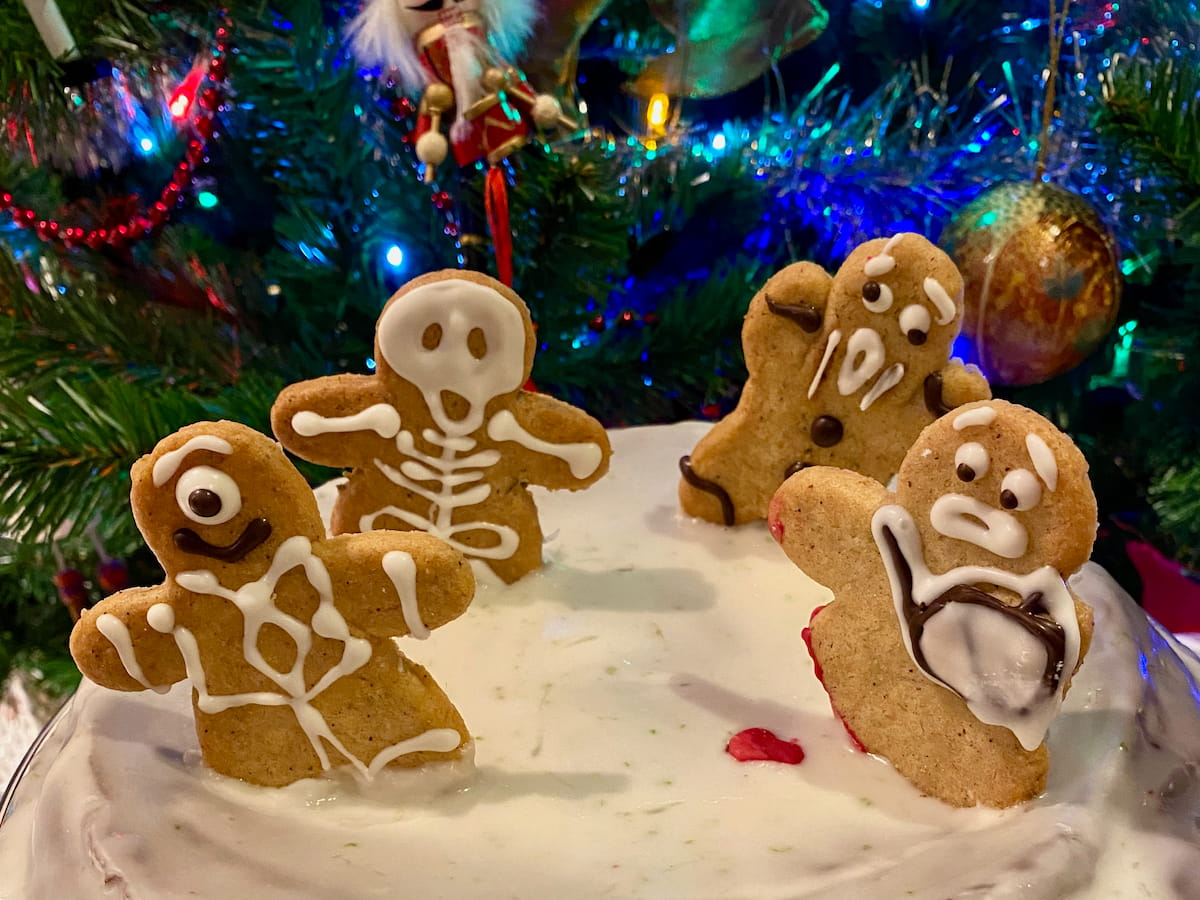 a white iced cake topped with halloween gingerbread cookies decorated as a skeleton, one-eyed monster and one with a severed leg