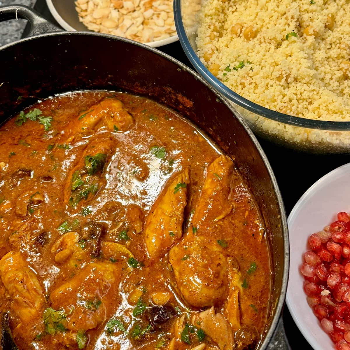 large dish with chicken tagine and prunes next to a bowl of cooked semolina, a pan of toasted almonds and pomegranate seeds