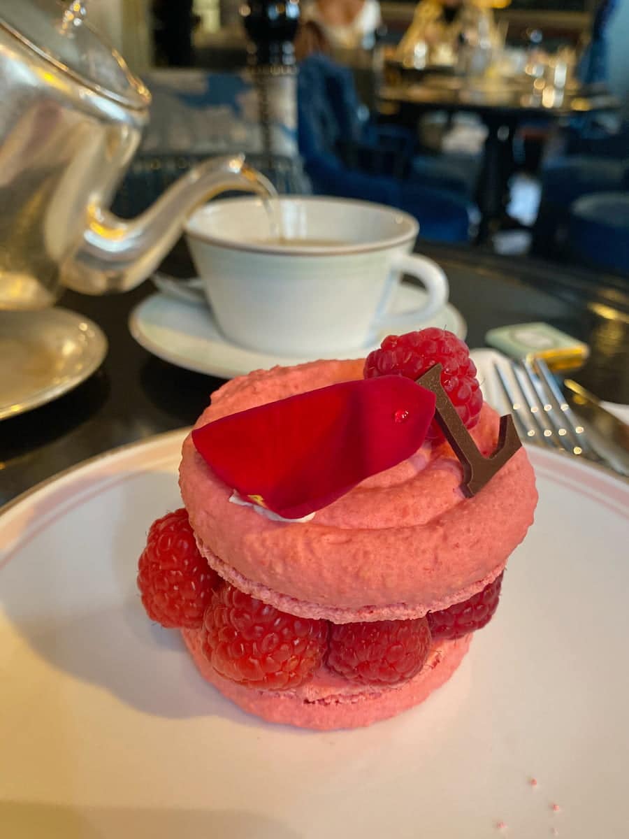 raspberries in a rose macaron dessert served on a plate in front of tea being poured