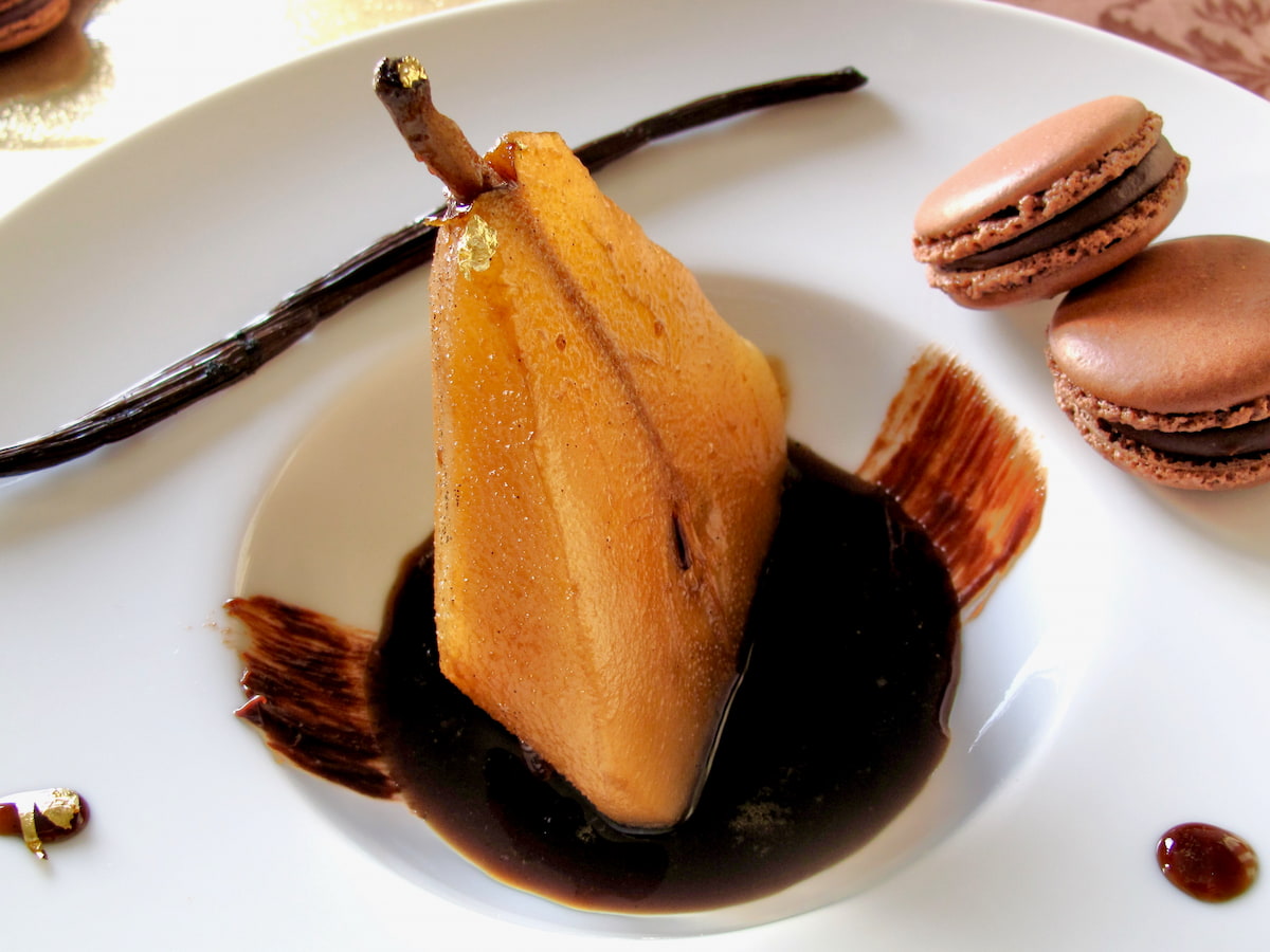half a poached pear sitting in a pool of coffee syrup in an elegant porcelain dish with a vanilla pod and chocolate macarons on the side
