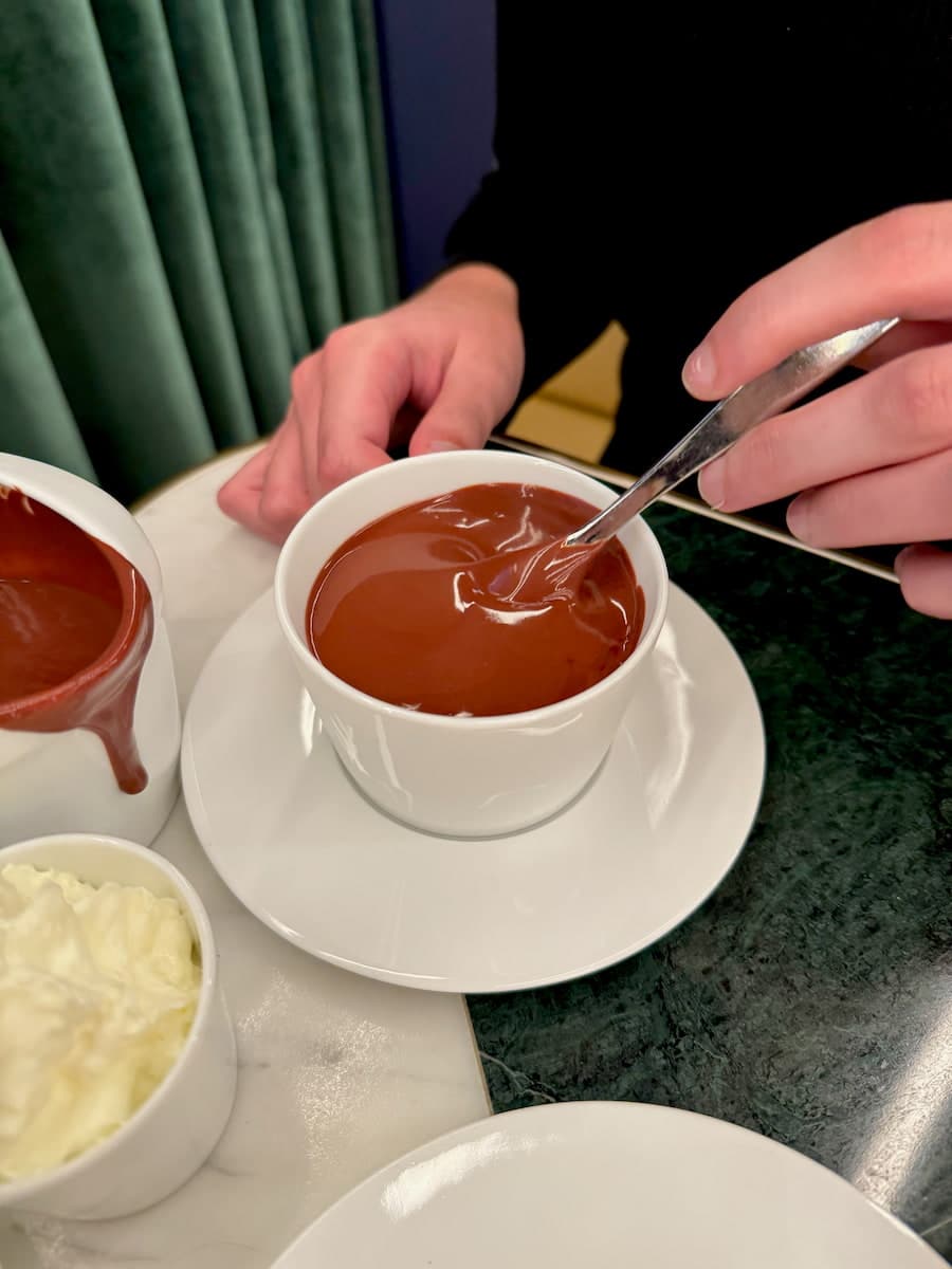 stirring an extremely thick dark hot chocolate in a cup next to a bowl of whipped cream