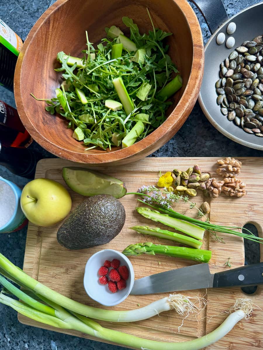 wooden salad bowl with arugula, avocado fand chopping board with spring onions, asparagus, apple and toasted pumpkin seeds