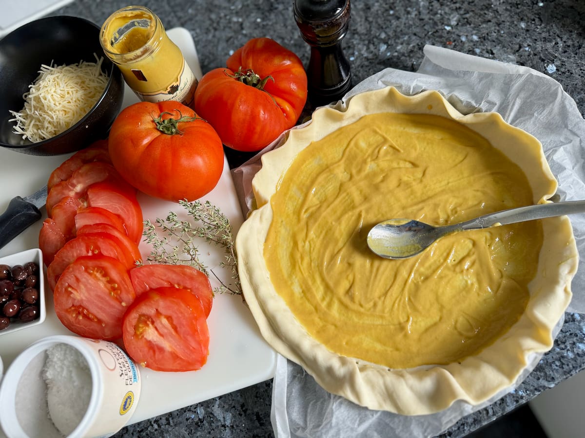 puff pastry in a tart pan, covered in a layer of Dijon mustard next to sliced tomatoes on a chopping board