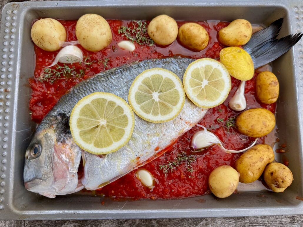 whole sea bream fish in a roasting tin topped with lemon slices and surrounded in tomato sauce, garlic and potatoes