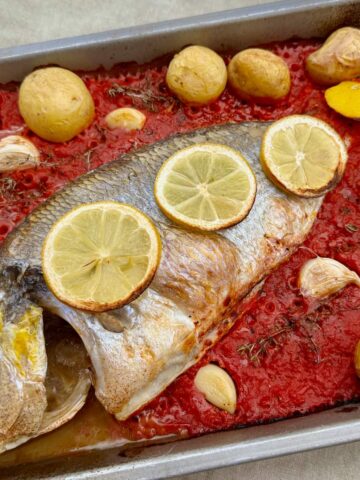 whole sea bream fish in a baking dish topped with tomato sauce, roast garlic cloves, lemon slices and potatoes