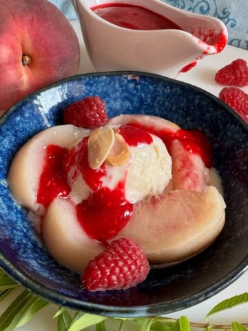shallow bowl of white poached vanilla peaches topped with homemade raspberry puree and vanilla ice cream with some fresh lemon verbena leaves