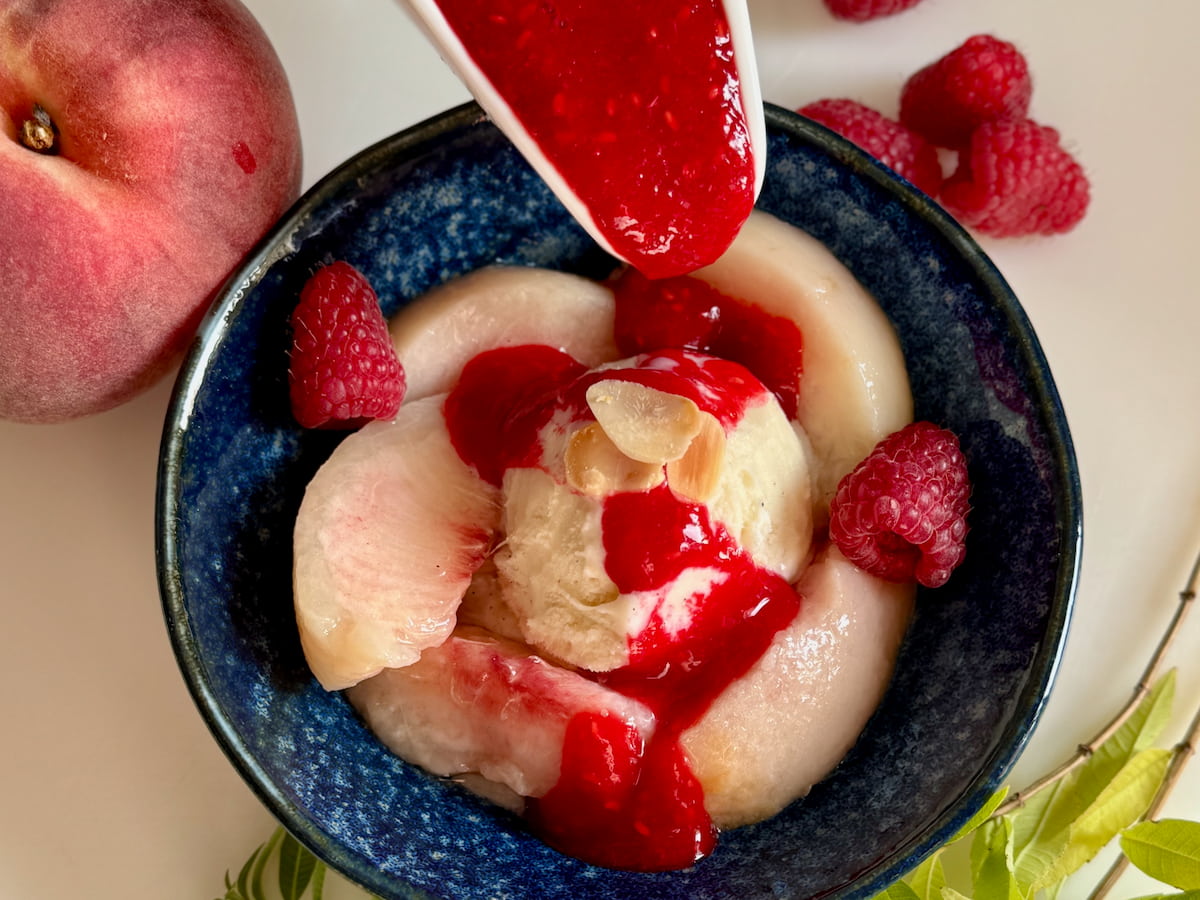 pouring homemade raspberry sauce onto poached peaches and vanilla ice cream in an individual dessert bowl
