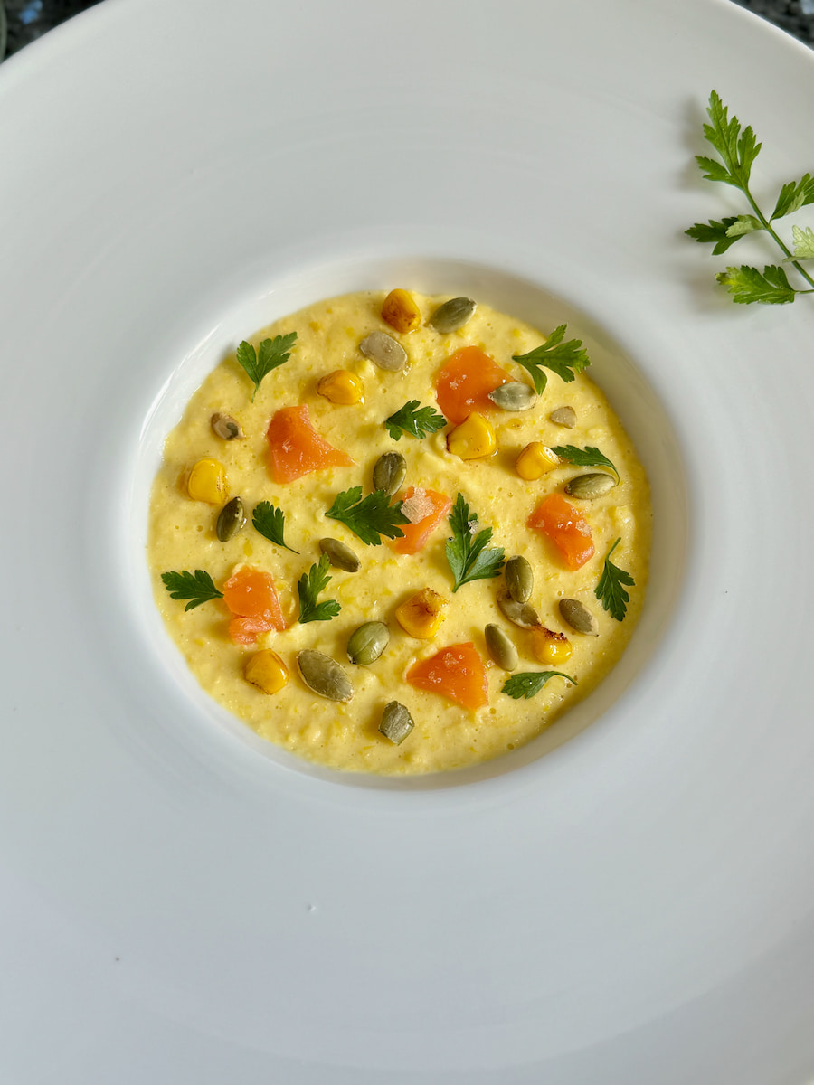 corn soup served in a large-rimmed porcelain bowl, topped with tiny herb leaves, pumpkin seeds, bits of smoked salmon and fried corn kernels