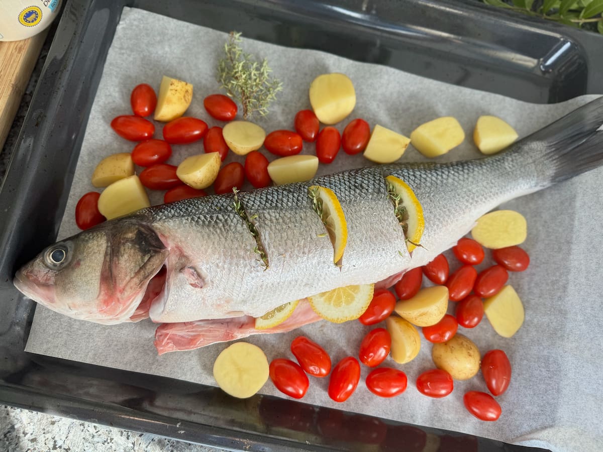 whole fish on a roasting tin with baking parchment and skin scored, stuffed with lemon, herbs and surrounded with potatoes and cherry  tomatoes