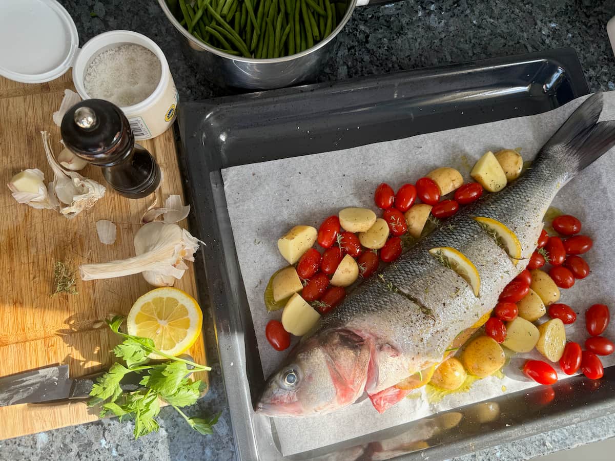 sea bass fish laid whole on a baking dish with potatoes, cherry tomatoes and herbs