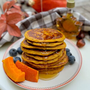 stack of orange hued cottage cheese pancakes with pumpkin purée with dribbling maple syrup on a plate with blueberries