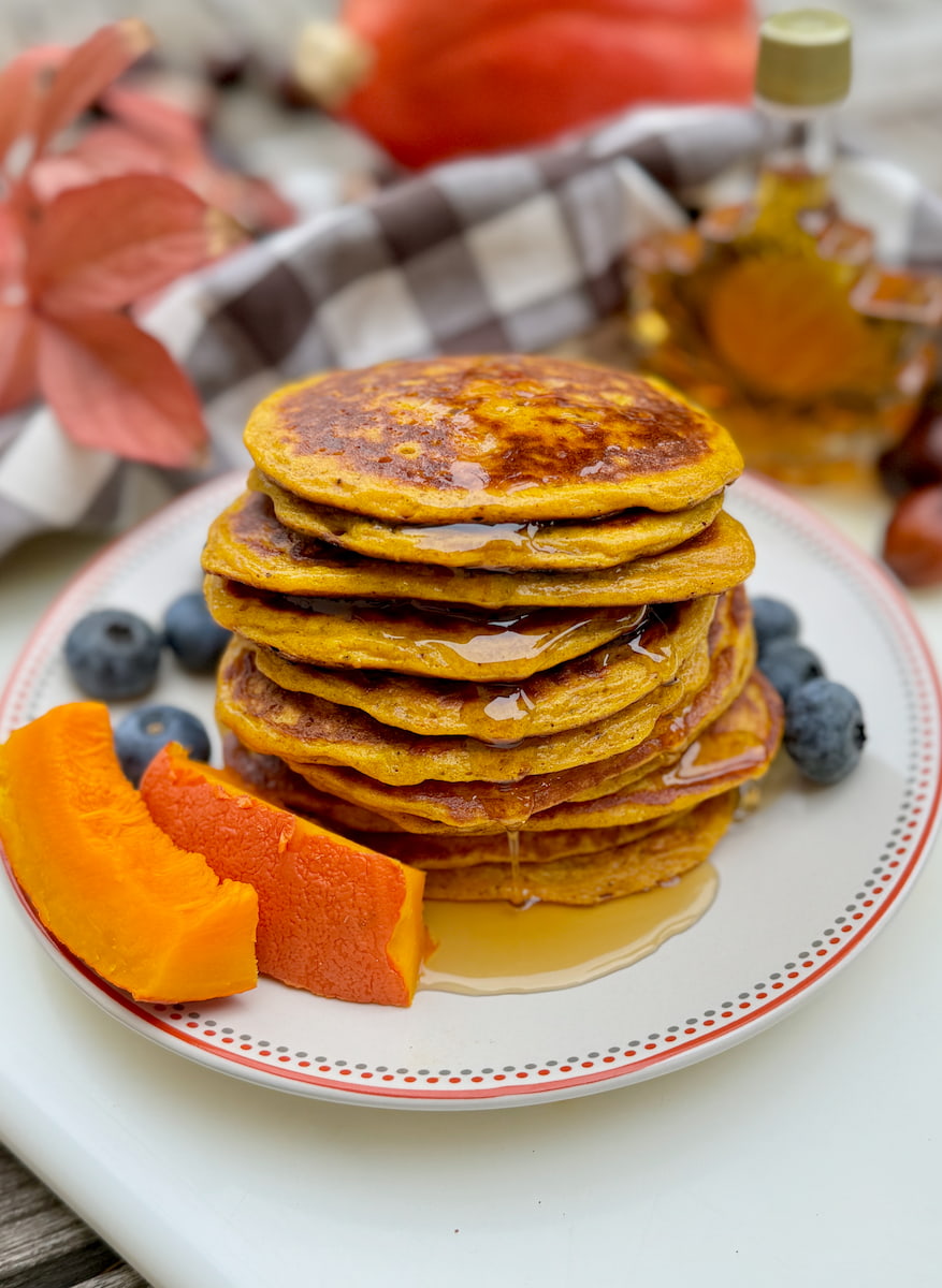 stack of cottage cheese pancakes being topped in original maple syrup next to blueberries and pumpkin