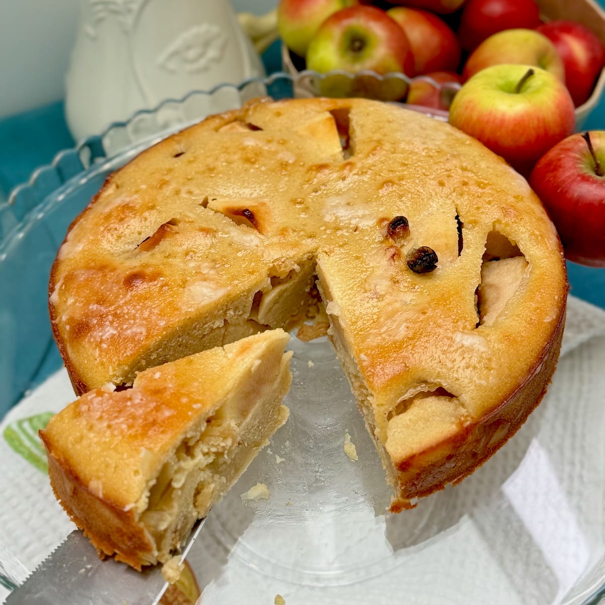 large apple cake showing moist inside with a slice removed, topped with a shiny glaze on a plate next to apples