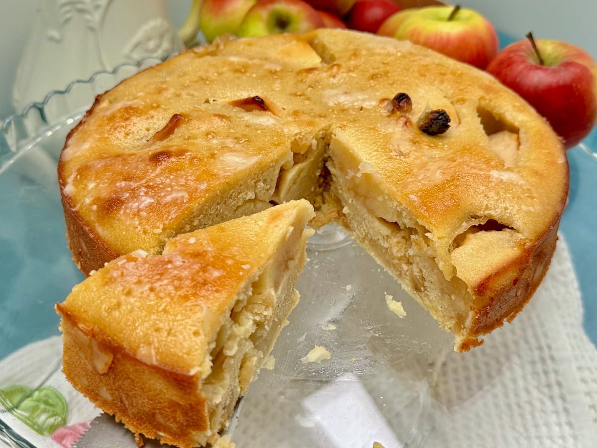 slice of moist cake filled with baked apples and raisins and topped with a shiny Calvados glaze in front of a whole cake and apples