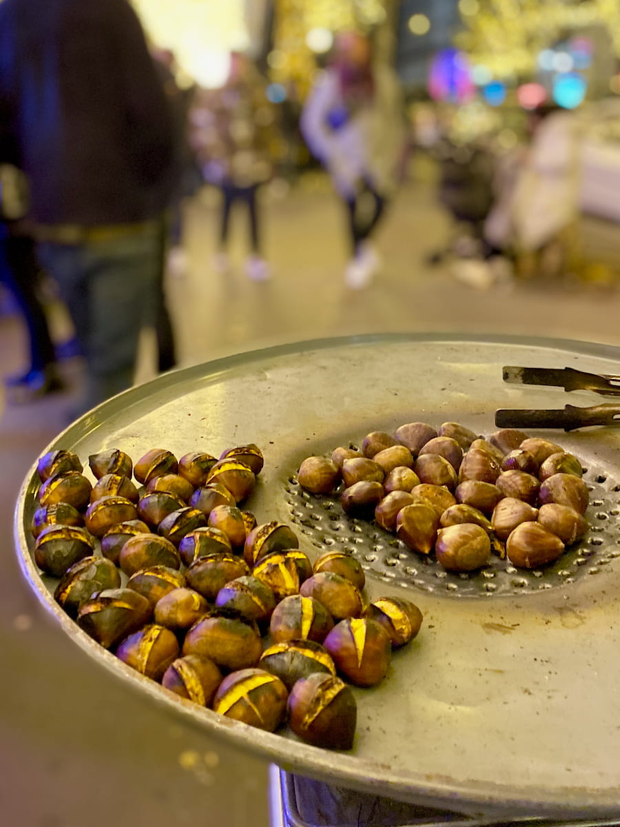 roasting chestnuts on a large tray next to the metro in Paris with Christmas lights in the background