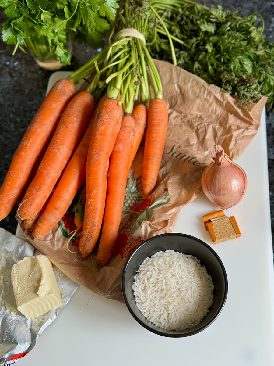 ingredients laid out to make carrot soup with a bunch of fresh carrots with their stalks, a small onion, butter, rice, stock and parsley