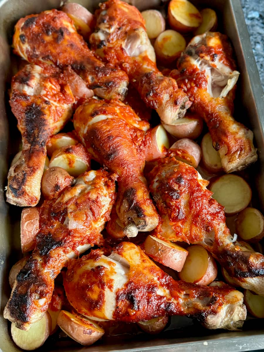 baking tray filled with roasting chicken drumsticks and potato slices covered in a BBQ marinade