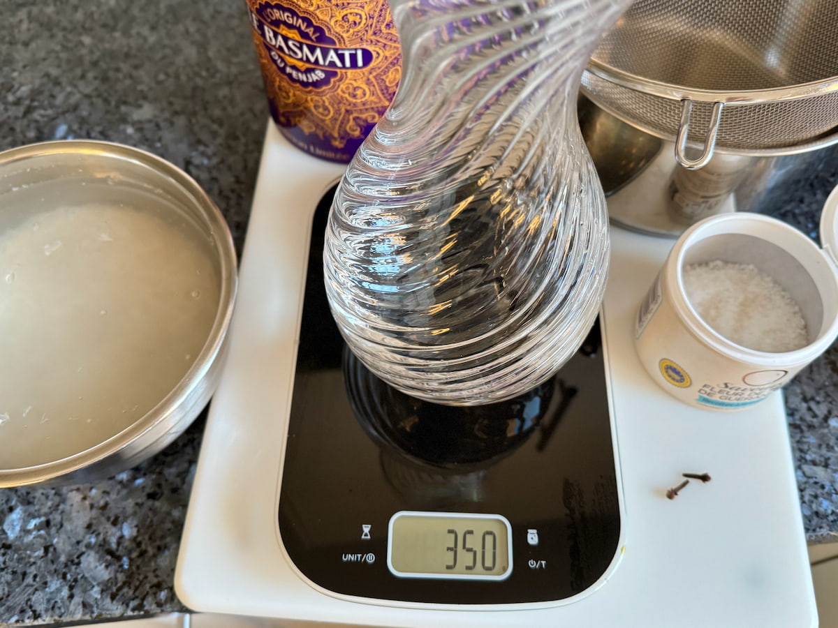 jug of water being weighed to 350 grams on digital scales next to salt, 2 cloves and a bowl of Basmati rice in water