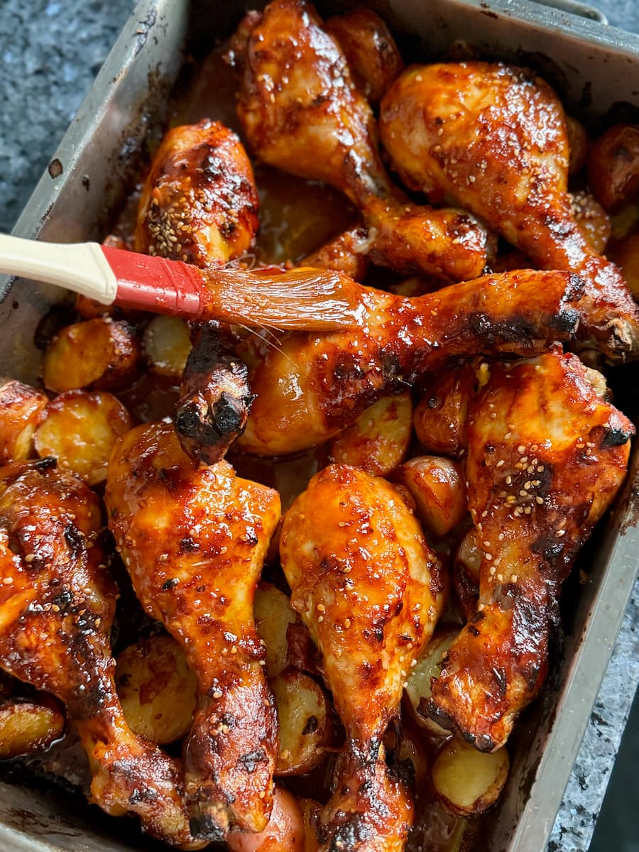 glazing over roasting chicken drumsticks and potatoes with a brush to coat them in sauce