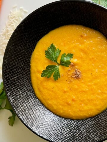 black bowl with a thick and creamy carrot soup, thickened with rice and topped with a sprig of fresh parsley and pinch of ground coriander