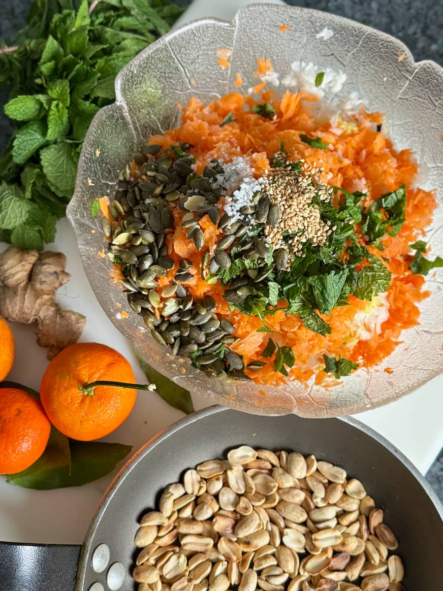 bowl with shredded carrots, ginger, fresh mint, flower salt, toasted pumpkin and sesame seeds and a pan of toasted peanuts with orange
