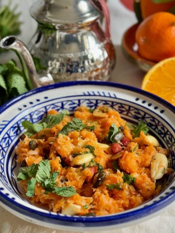 Moroccan handpainted bowl filled with a raw carrot salad topped with fresh mint, toasted nuts and seeds next to oranges and a Moroccan teapot