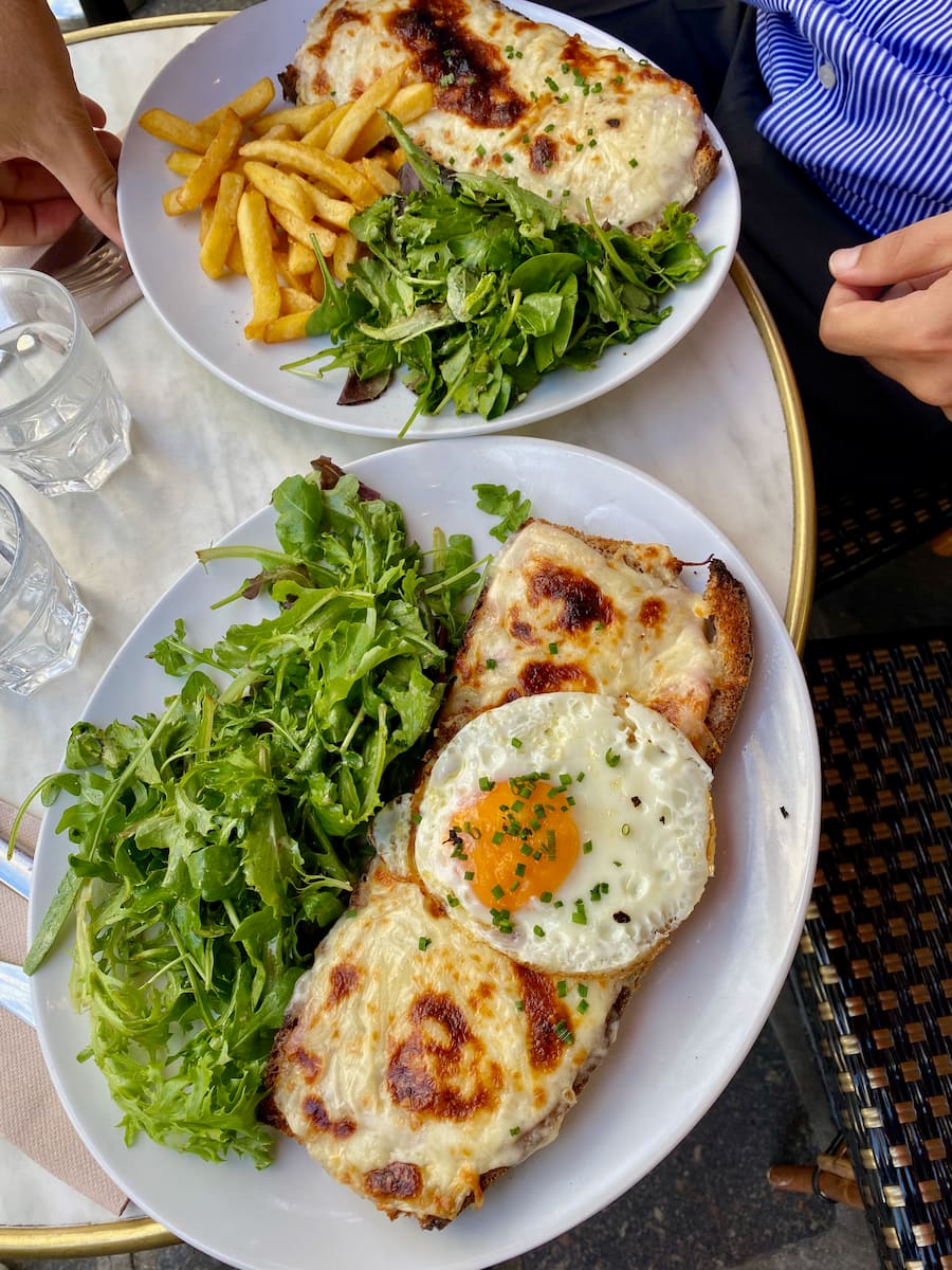 two plates showing a Croque Monsieur with salad and fries and a Croque Madame with a fried egg on top, served with green salad in a Parisian bistro