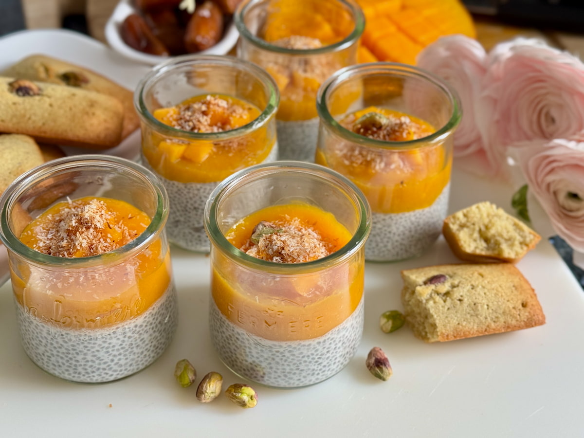 glass pots with creamy coconut chia pudding topped with mango sauce, pistachios and toasted coconut next to pistachio financier cakes
