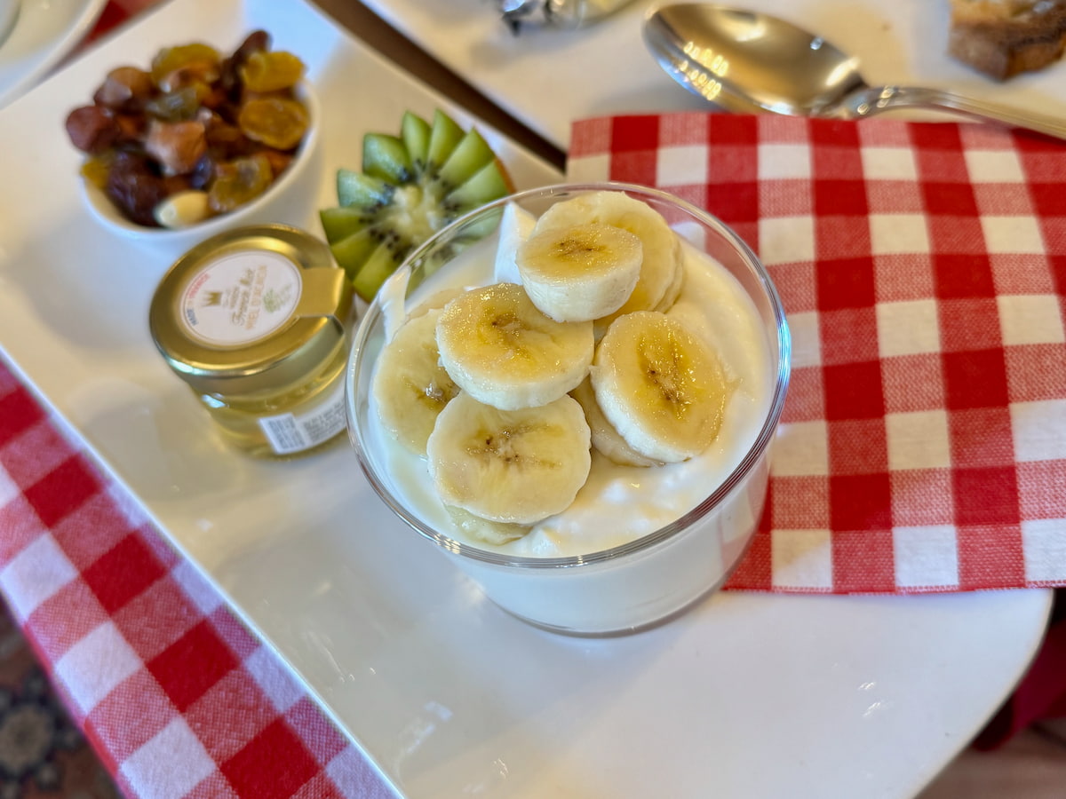 sliced banana on top of a glass bowl of fromage blanc with a pot of honey, nuts and dried fruits and a kiwi