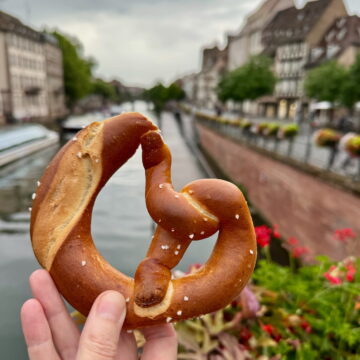 holding a salty bretzel in Strasbourg in front of the river with flowers and timbered buildings in the background