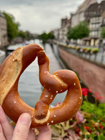 holding a salty bretzel in Strasbourg in front of the river with flowers and timbered buildings in the background
